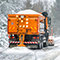 An orange snowplow driving along a snow-swept highway lined with forest trees during a snowstorm