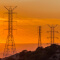 A row of pylons strung with long power lines on a brush-covered hillside silhouetted against a vivid orange sunset