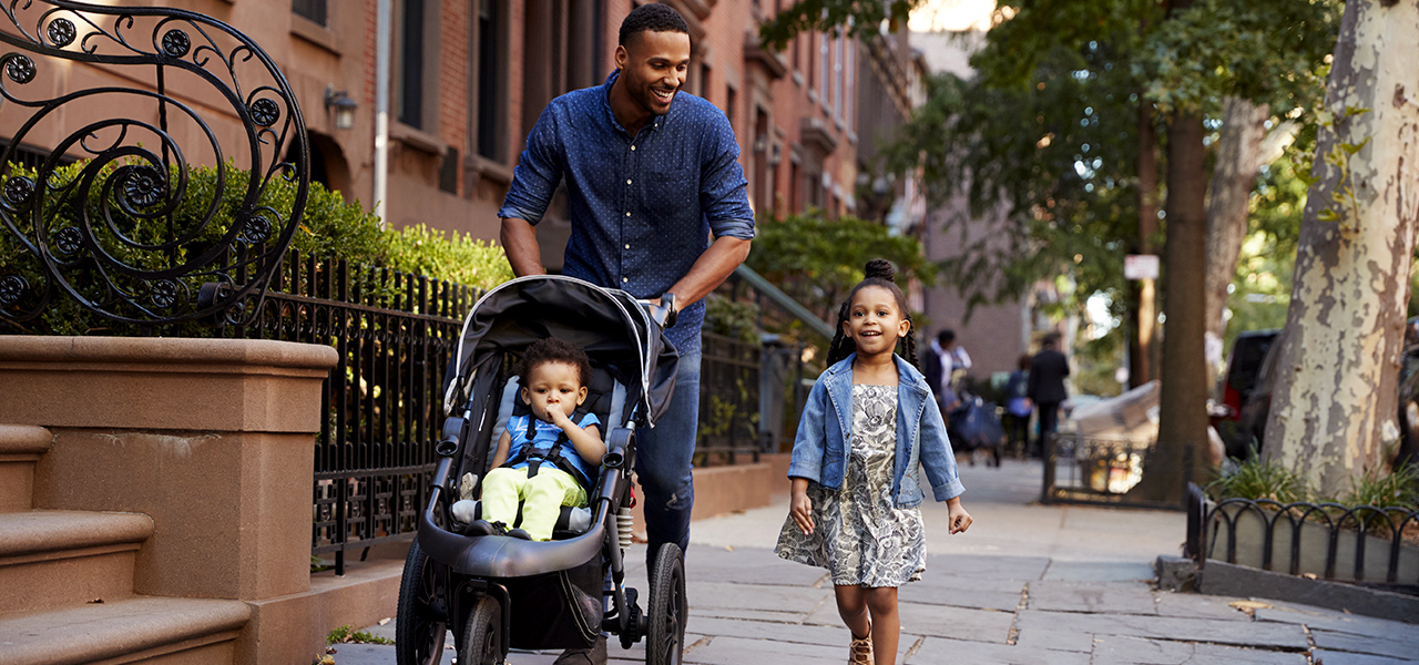 Man pushing a stroller on a city sidewalk and a child walking beside him